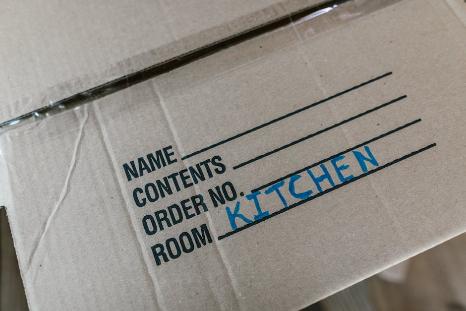 Two women engaged in a home relocation process are standing among several large cardboard moving boxes, some sealed with red tape, in an indoor setting with a textured grey wall background. One woman,